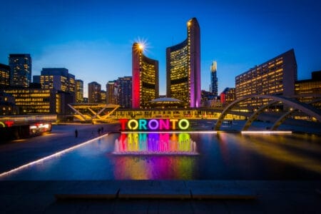 Nathan Phillips Square in Toronto am Abend. Im Zentrum steht ein großes, bunt beleuchtetes „TORONTO“-Schriftzug vor einem Springbrunnen. Dahinter ragen moderne Hochhäuser empor, darunter die markant geschwungenen Türme des modernen Rathauses von Toronto. Der Himmel ist klar und geht allmählich vom Tageslicht in die Nacht über.
