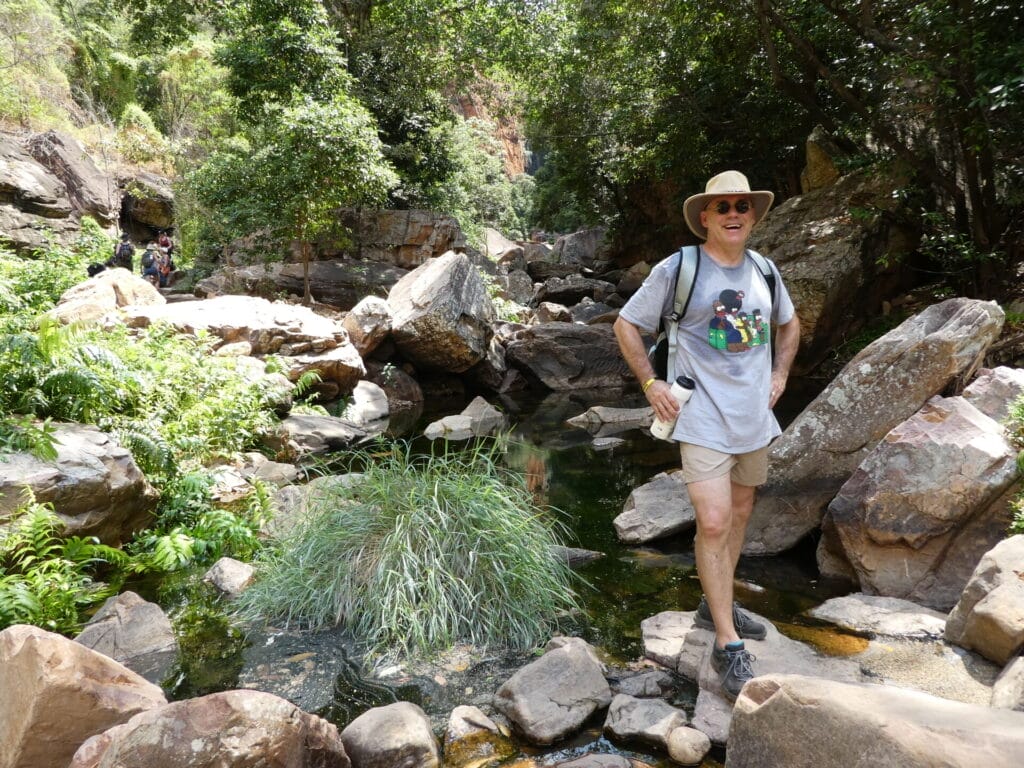 „Person steht mit Rucksack auf Felsen in einer grünen Schlucht mit Bachlauf und umgebender Vegetation.“