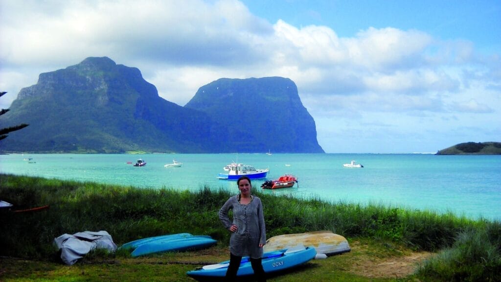„Person steht unter einer großen, geschwungenen Felsformation an einer Küstenklippe mit Blick auf das Meer und hält sich mit einer Hand am Fels fest.“