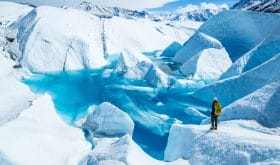 Standing near the edge of large blue pool on top of the Matanuska Glacier. A young woman holding an ice axe with a backpack and helmet looks out over the lake.