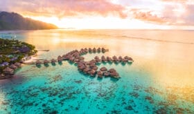 Luxury travel vacation aerial of overwater bungalows resort in coral reef lagoon ocean by beach. View from above at sunset of paradise getaway Moorea, French Polynesia, Tahiti, South Pacific Ocean.