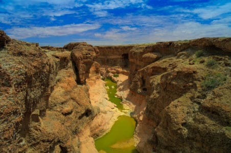 „Sesriem Canyon im Namib-Naukluft-Park, Namibia – tiefe Schlucht mit braunen Felswänden und grünem Fluss unter blauem Himmel mit Wolken.“
