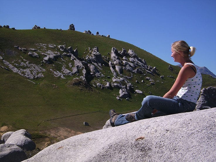 „Person sitzt auf einem großen Felsen und blickt über eine hügelige Landschaft mit markanten grauen Felsformationen unter klarem blauem Himmel.“