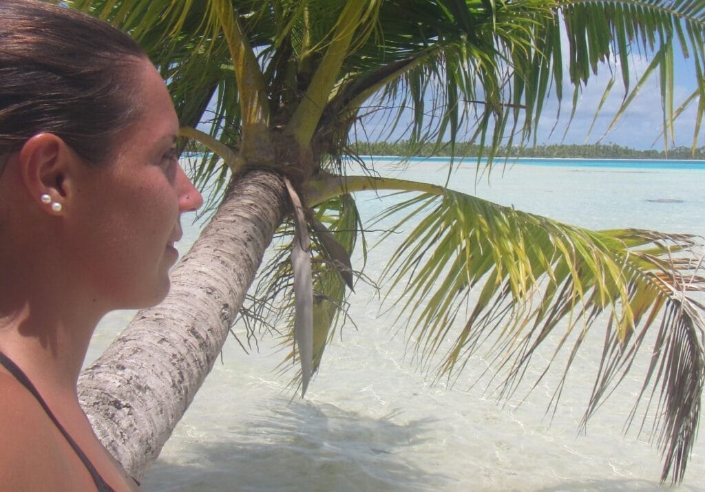 „Person steht im seichten, türkisfarbenen Wasser nahe einer gebogenen Palme, mit klarem Lagunenwasser und einer palmengesäumten Insel im Hintergrund.“