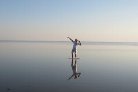 Ein Mann mit weißem T-Shirt und Shorts steht auf einer spiegelglatten Wasseroberfläche, die den wolkenlosen Himmel und die Person reflektiert.