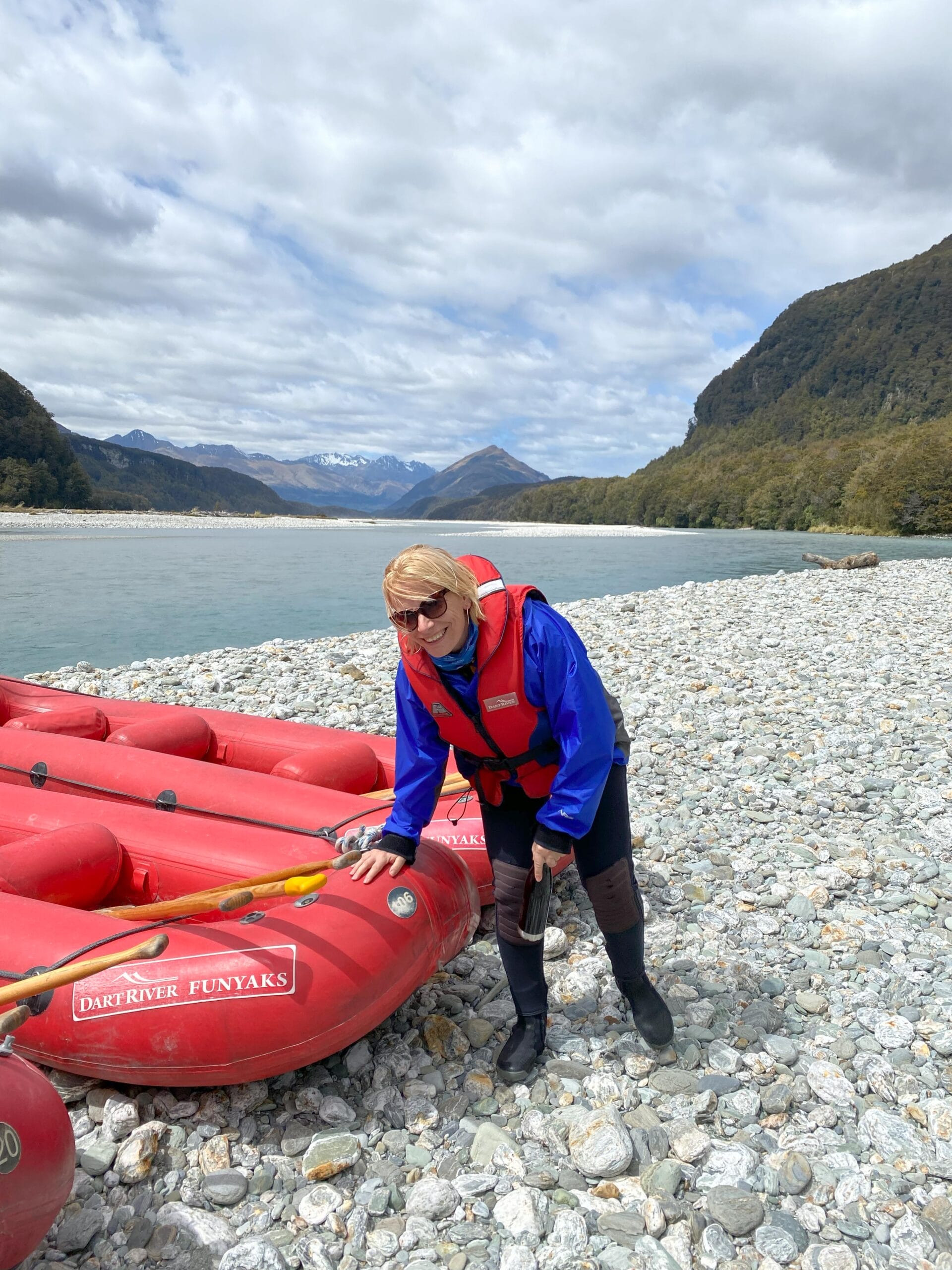 Person bit blauer Kacke und roter Schwimmweset an einem roten Schlauchboot für Dart River Funyaks am Kiesufer in Neuseeland mit Bergen und Fluss im Hintergrund.