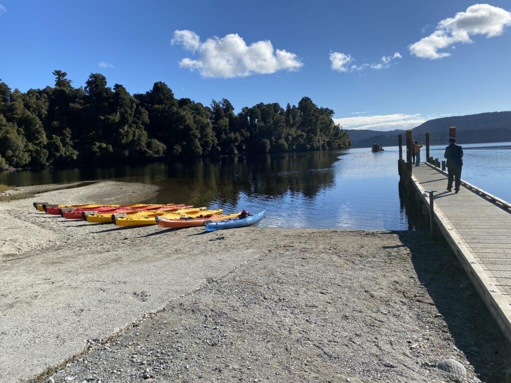 Bunte Kajaks am Kiesstrand eines klaren Sees in Neuseeland mit Holzsteg und bewaldeten Hügeln im Hintergrund.