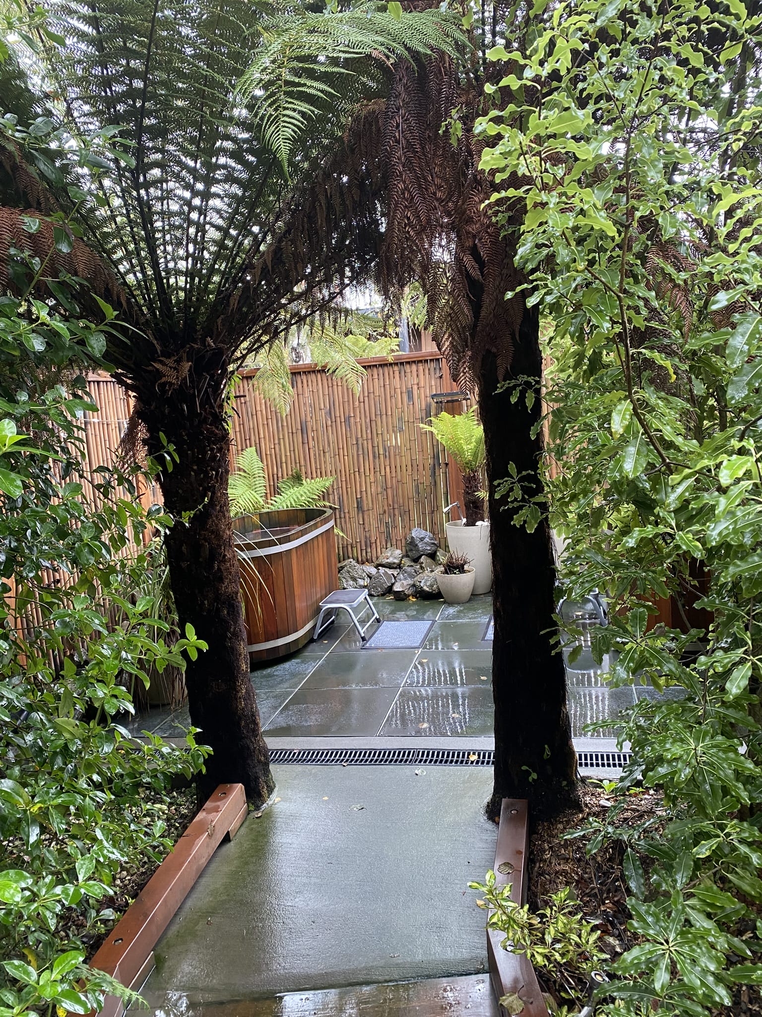 Privater Outdoor-Whirlpool auf Terrasse in Neuseeland, umgeben von Bambuswänden, Farnbäumen und üppiger Vegetation.