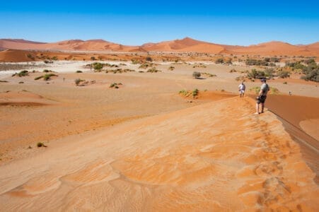 Personen stehen auf einer roten Sanddüne in Sossusvlei, Namibia, mit Blick auf die weite Wüstenlandschaft unter blauem Himmel.