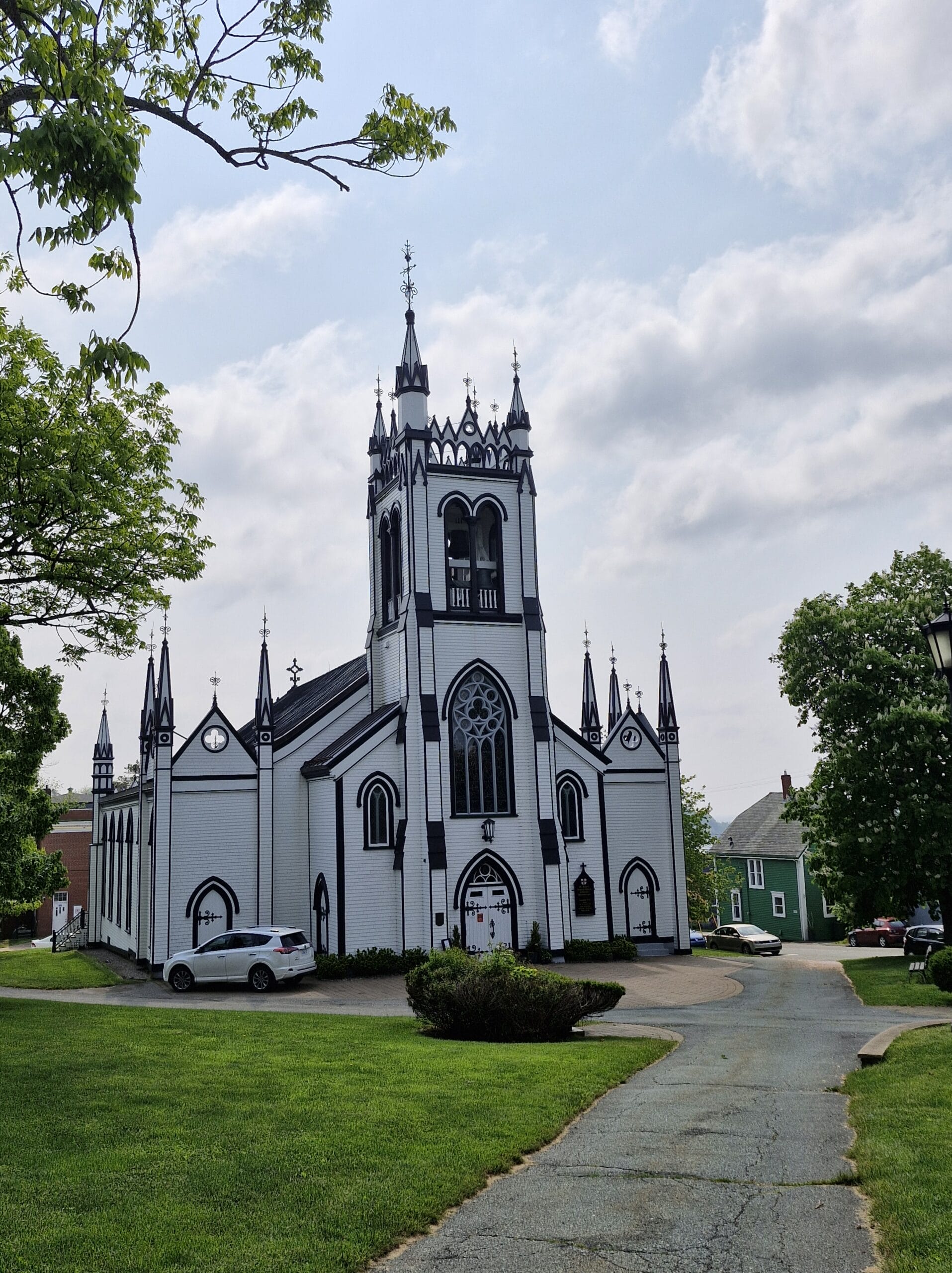„Weiß‑schwarze Kirche im neugotischen Stil mit turmartigem Eingang, spitzen Zierelementen und großen Spitzbogenfenstern, umgeben von Rasenflächen, Bäumen und einem Gehweg unter leicht bewölktem Himmel.“