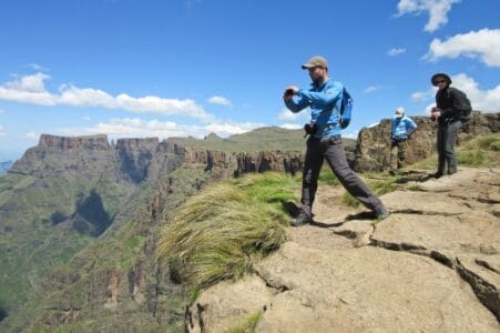 Mehrere Wanderer stehen auf einem felsigen Plateau mit spektakulärem Blick auf das Drakensberg-Amphitheater in Südafrika. Im Vordergrund ist eine Person in Outdoor-Bekleidung zu sehen, die ein Foto macht. Die Landschaft zeigt steile Felswände, grüne Täler und einen klaren blauen Himmel mit vereinzelten Wolken. Das Bild vermittelt Abenteuer, Naturerlebnis und Weitblick.